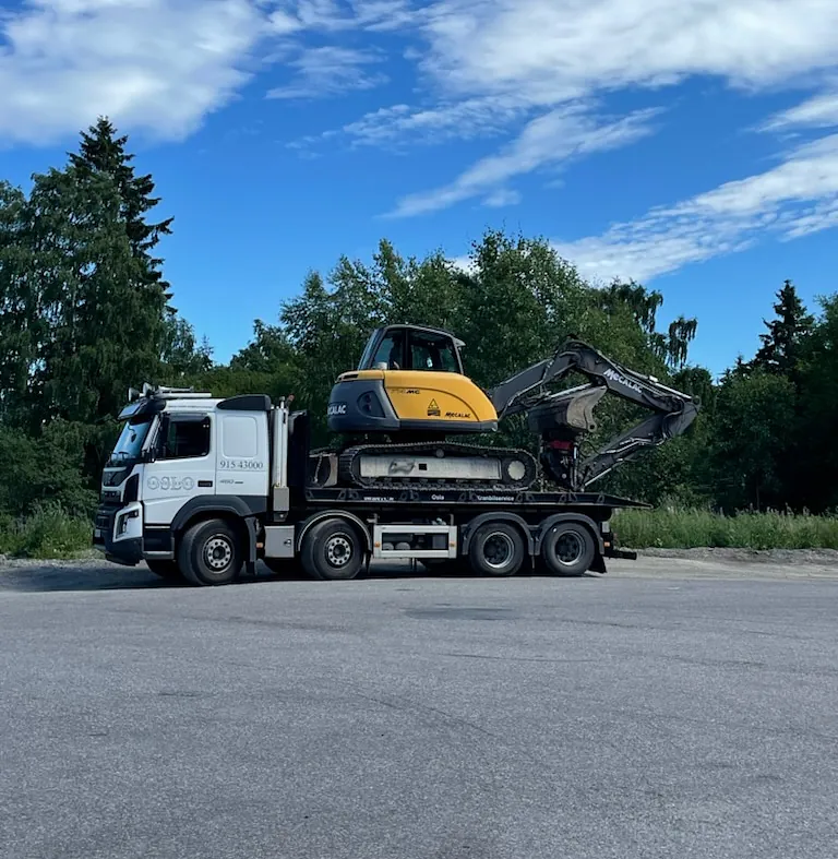 Hvit lastebil som frakter gul gravemaskin